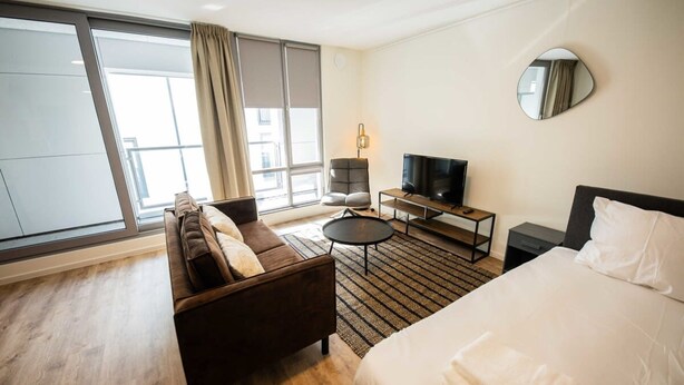 Habitación de hotel con cama, sofá, televisión, una mesita y ventana con cortinas.