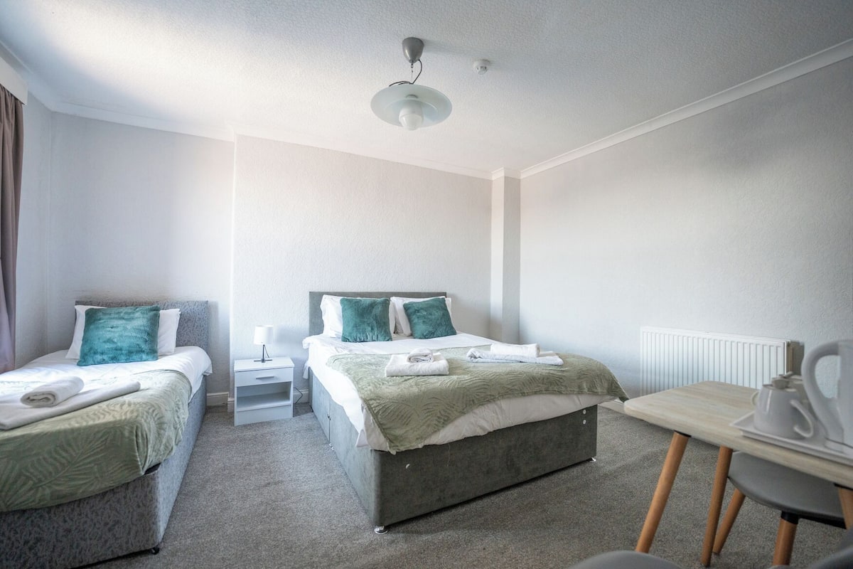 A hotel room with two single beds, a desk with a chair, a bedside table with a lamp, and a radiator.