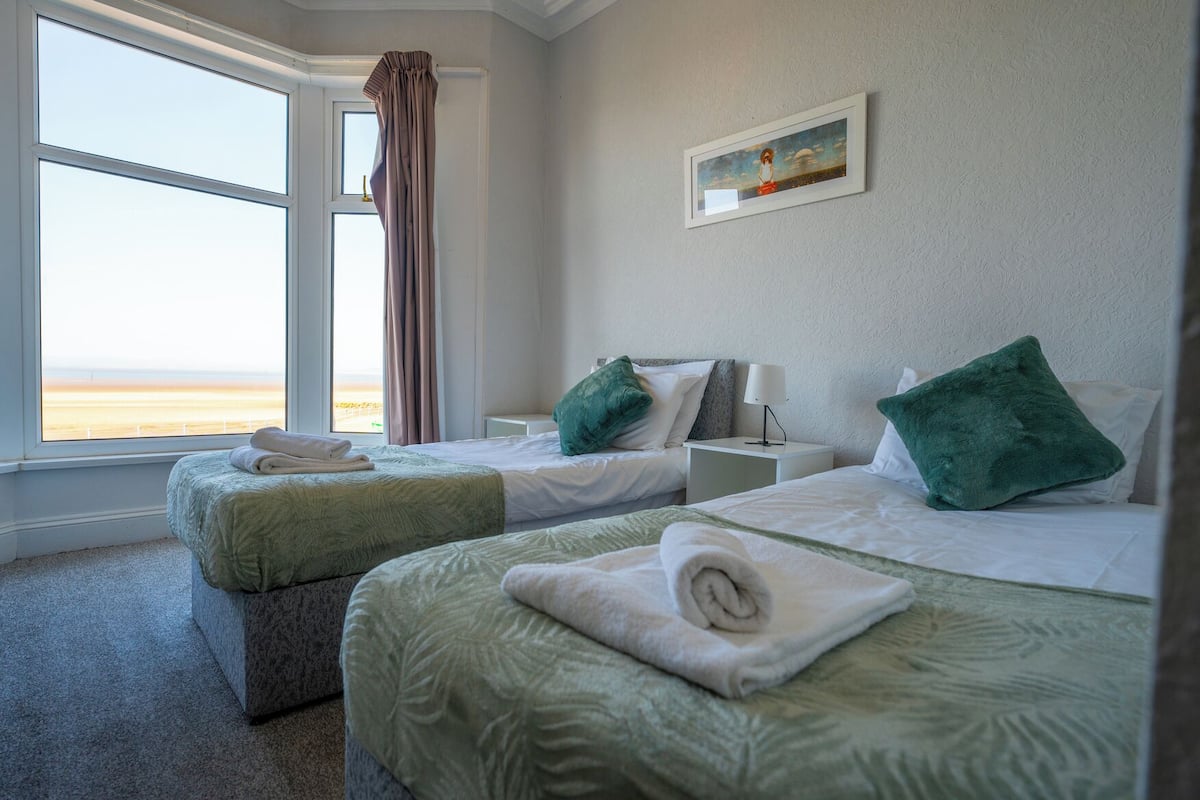 A hotel room with two beds, a large window with a view of the beach, a painting on the wall, and a nightstand with a lamp.