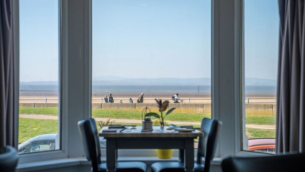 A dining area with a table and chairs, a view of a beach with people and a cyclist, and a clear sky. - The Clifton Hotel (Morecambe)