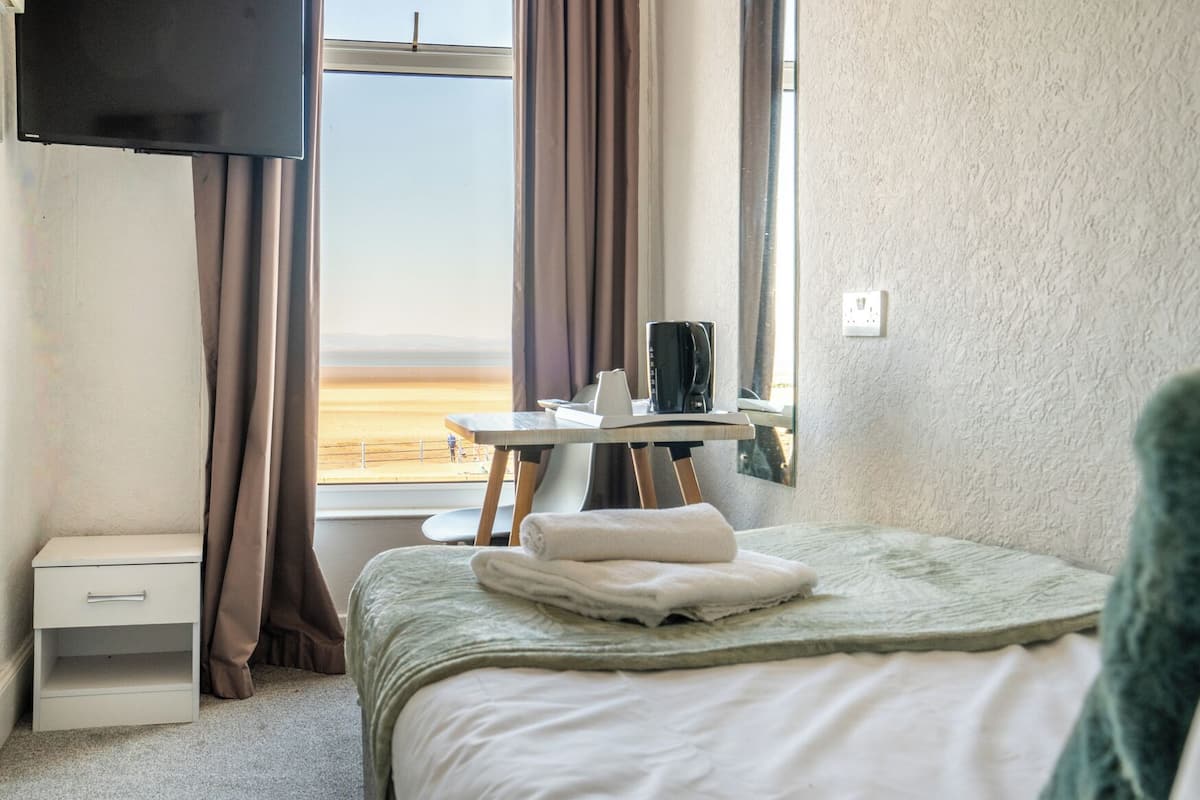 A hotel room with a bed, a small table with a coffee maker, a TV mounted on the wall, and a view of the beach through the window.