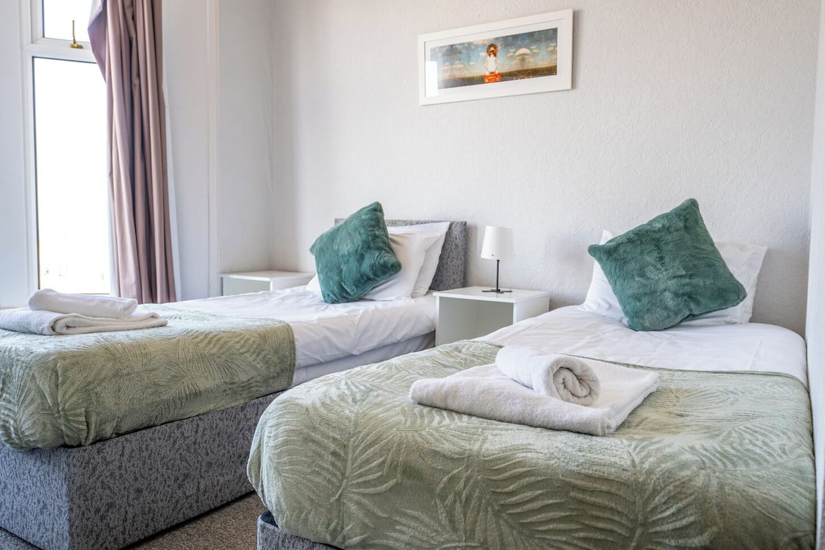 A hotel room with two beds, each with a green pillow and a white blanket. There is a painting on the wall above the beds.