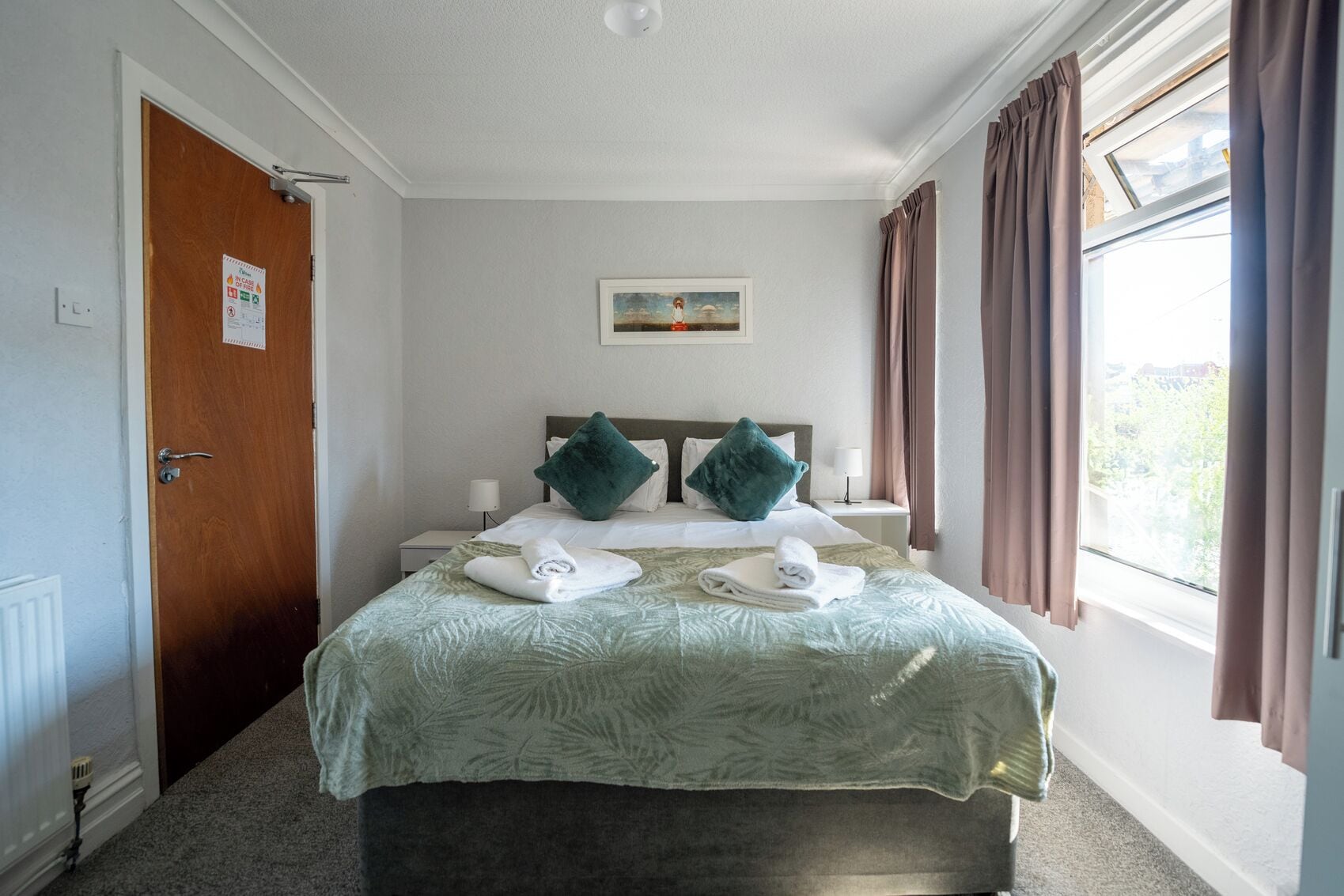 A hotel room with a bed, a painting on the wall, and a window with curtains.