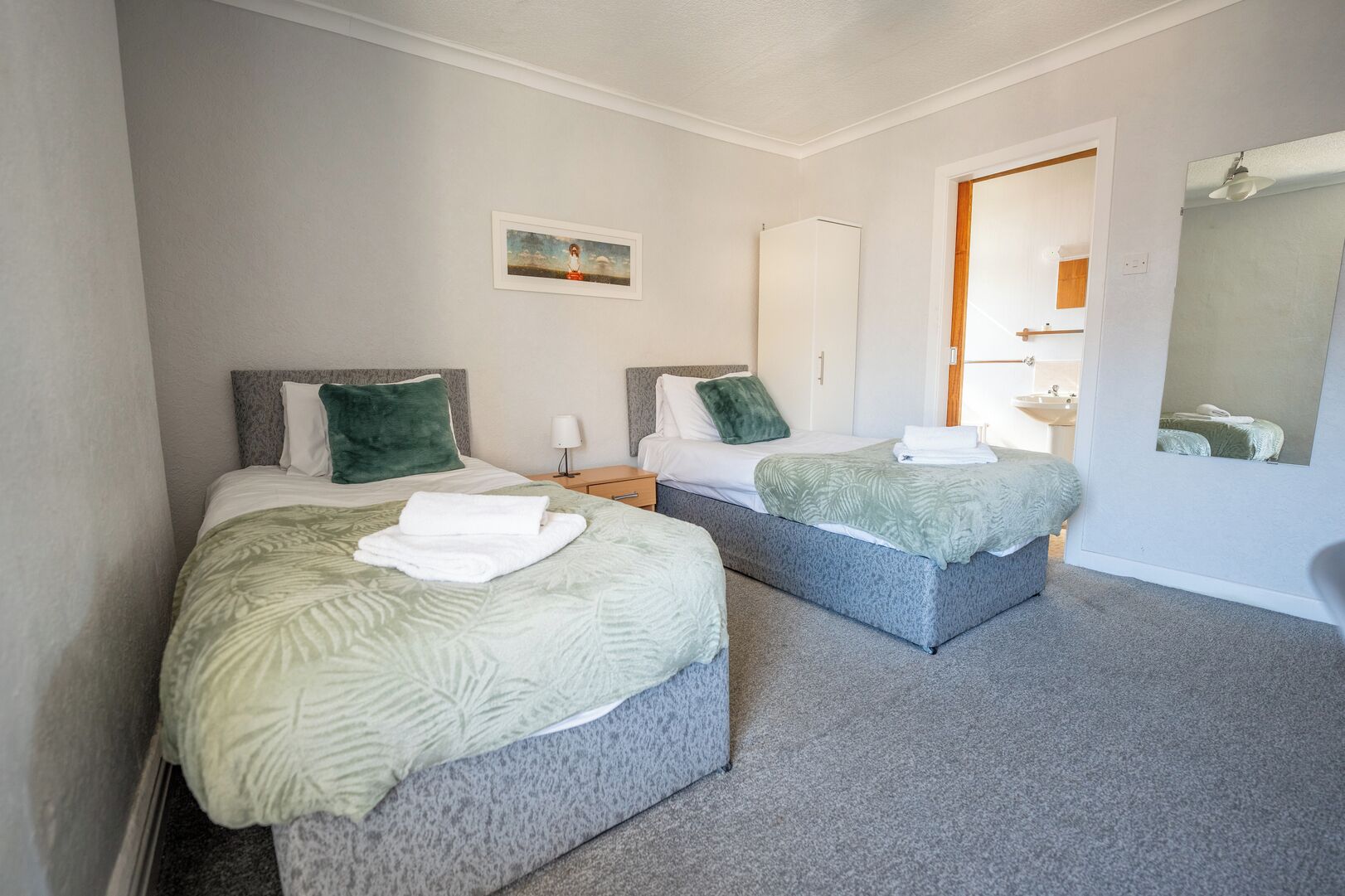 A hotel room with two beds, a small bedside table with a lamp, a wardrobe, and a mirror on the wall.