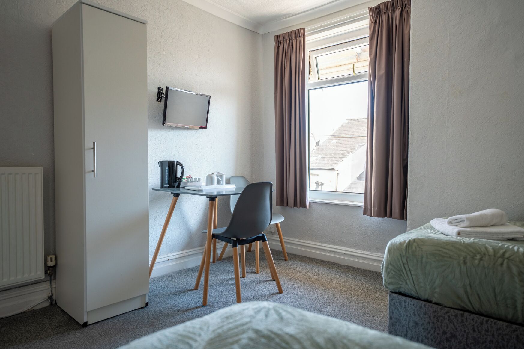 A hotel room with a bed, a desk with a chair, a radiator, a window with curtains, and a television mounted on the wall.