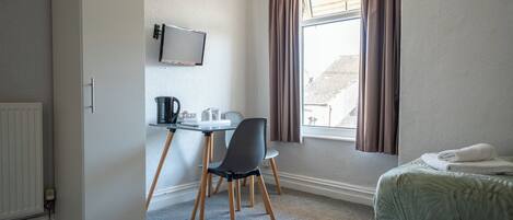 A hotel room with a bed, a desk with a chair, a radiator, a window with curtains, and a television mounted on the wall.