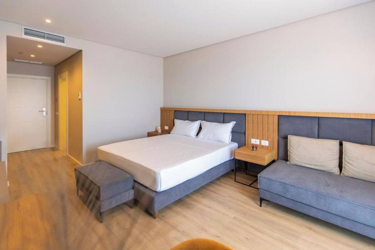 A modern hotel room with a large bed, a grey sofa, and a wooden nightstand.