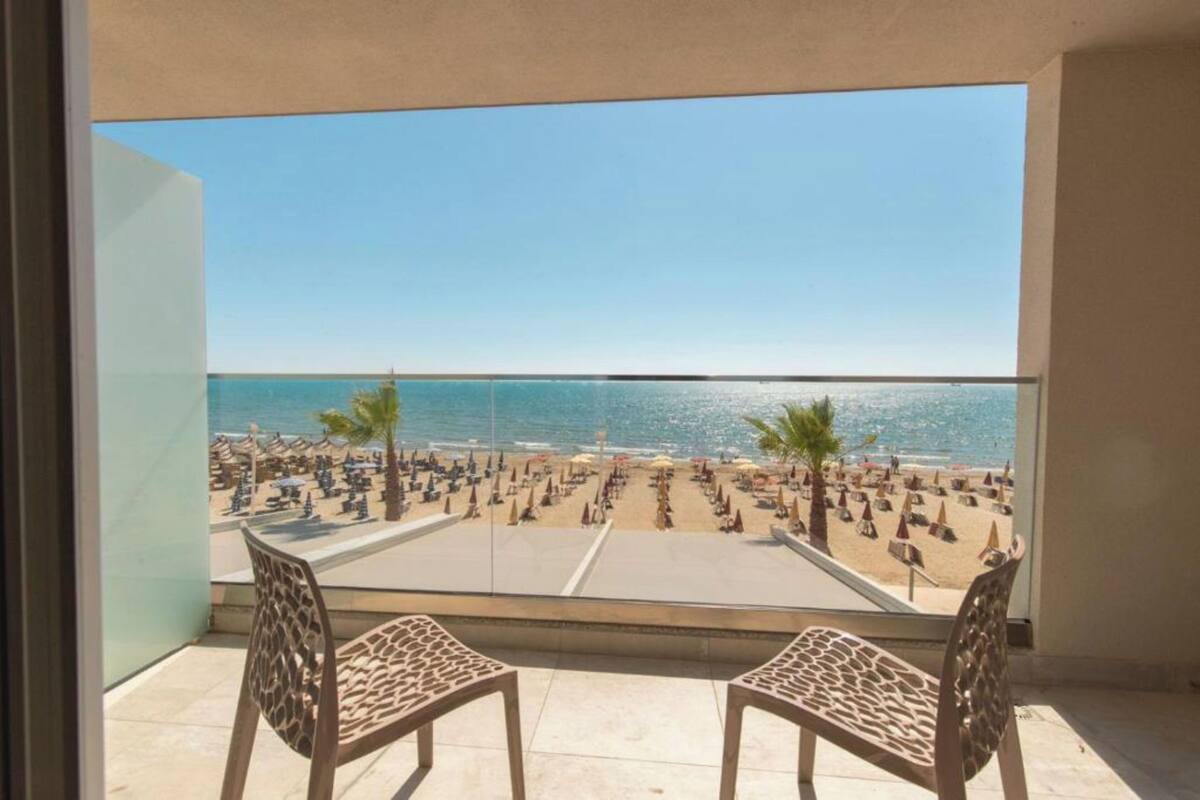 A balcony with two chairs, overlooking a beach with umbrellas and palm trees.