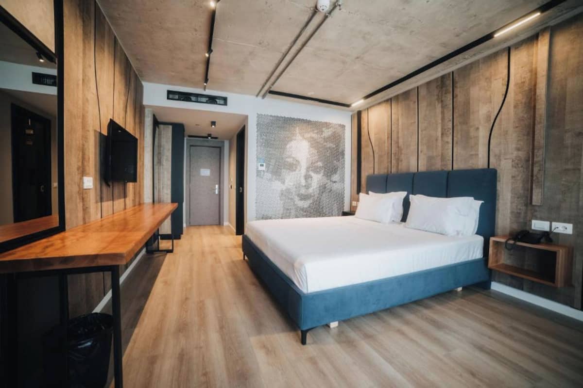 A modern hotel room with a wooden floor, a bed with a blue headboard, a desk, and a wall-mounted TV.