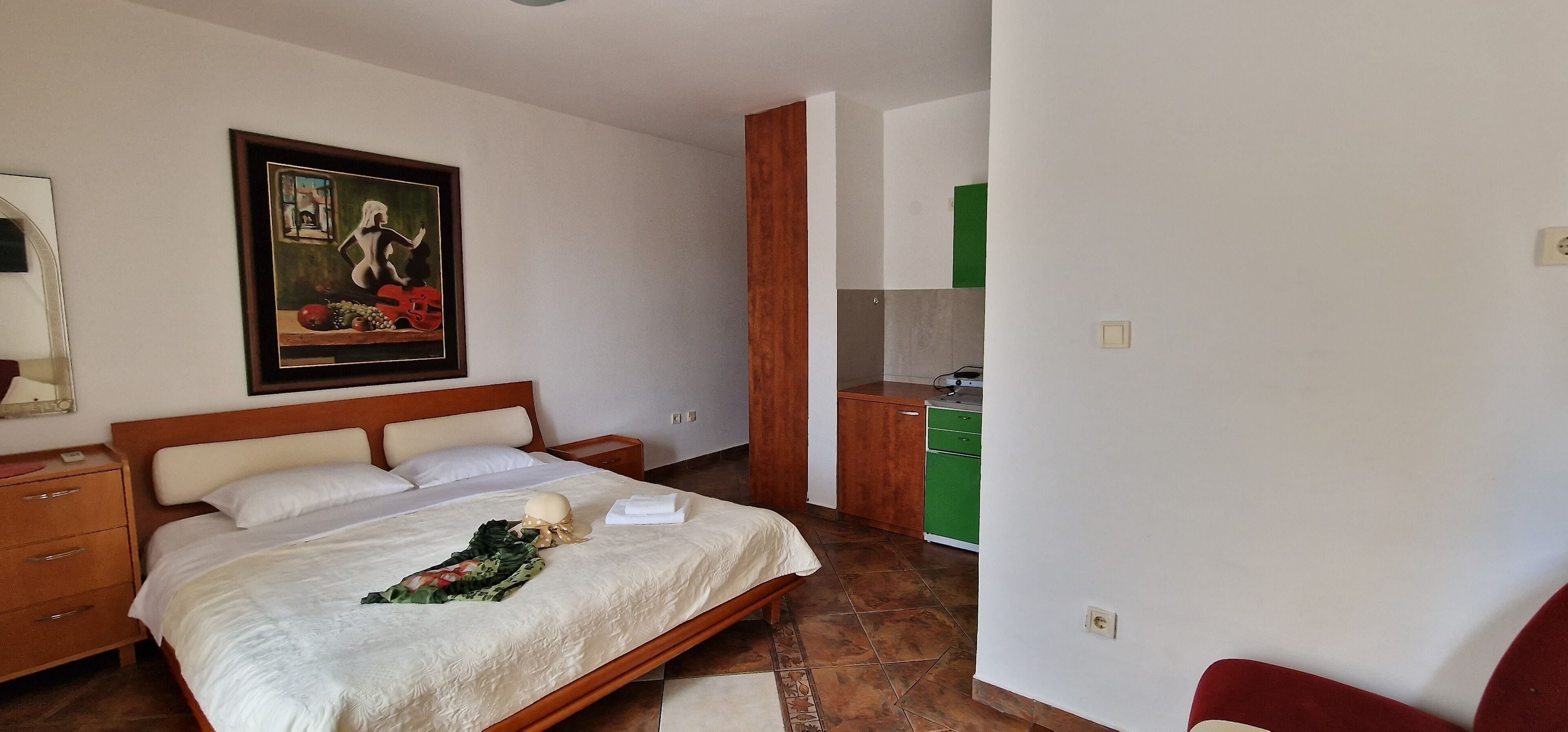 Une chambre d’hôtel comprenant un lit, une table de chevet, un tableau, une armoire en bois et une kitchenette.