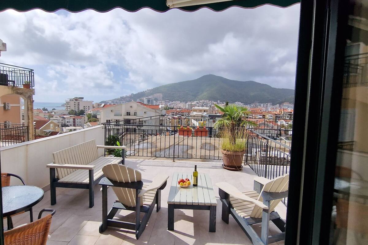 A balcony with outdoor furniture, a potted plant, and a view of a cityscape and mountains.