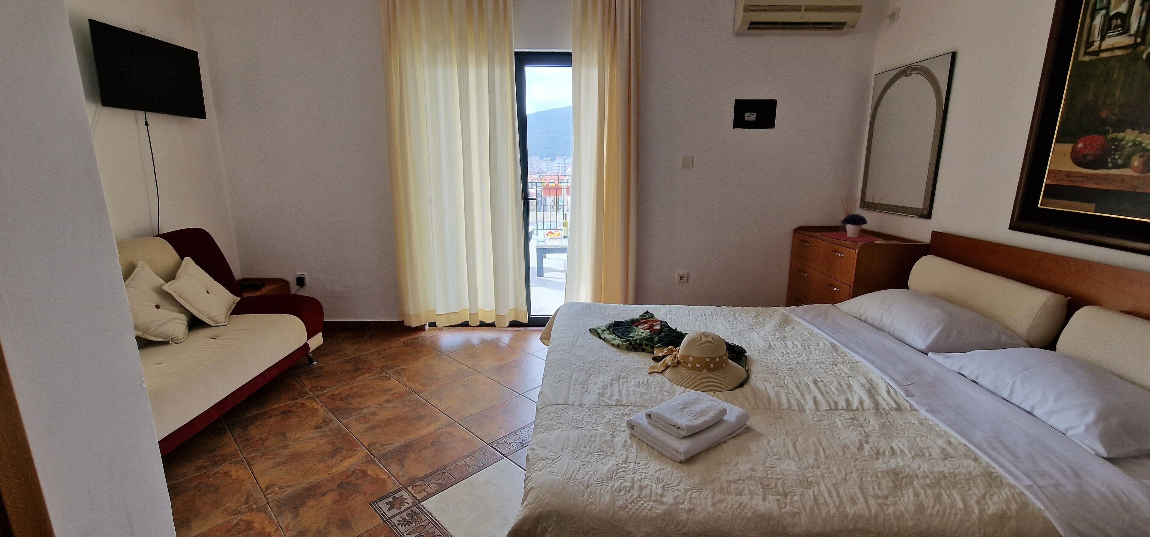 Une chambre d’hôtel avec un lit, un canapé, une télévision, une fenêtre avec des rideaux et un tableau au mur.