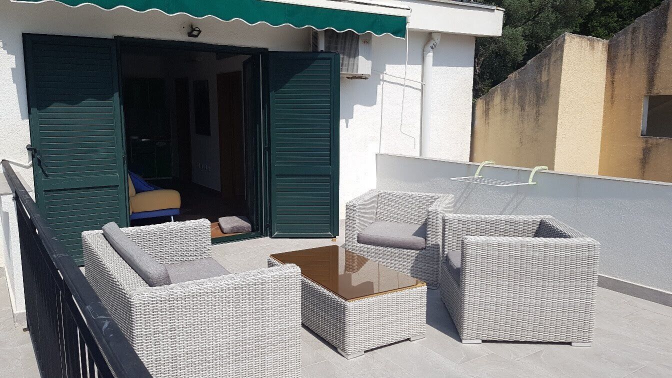 Un área de estar al aire libre con mobiliario de mimbre y una mesa con cubierta de vidrio, junto a un edificio con contraventanas verdes.