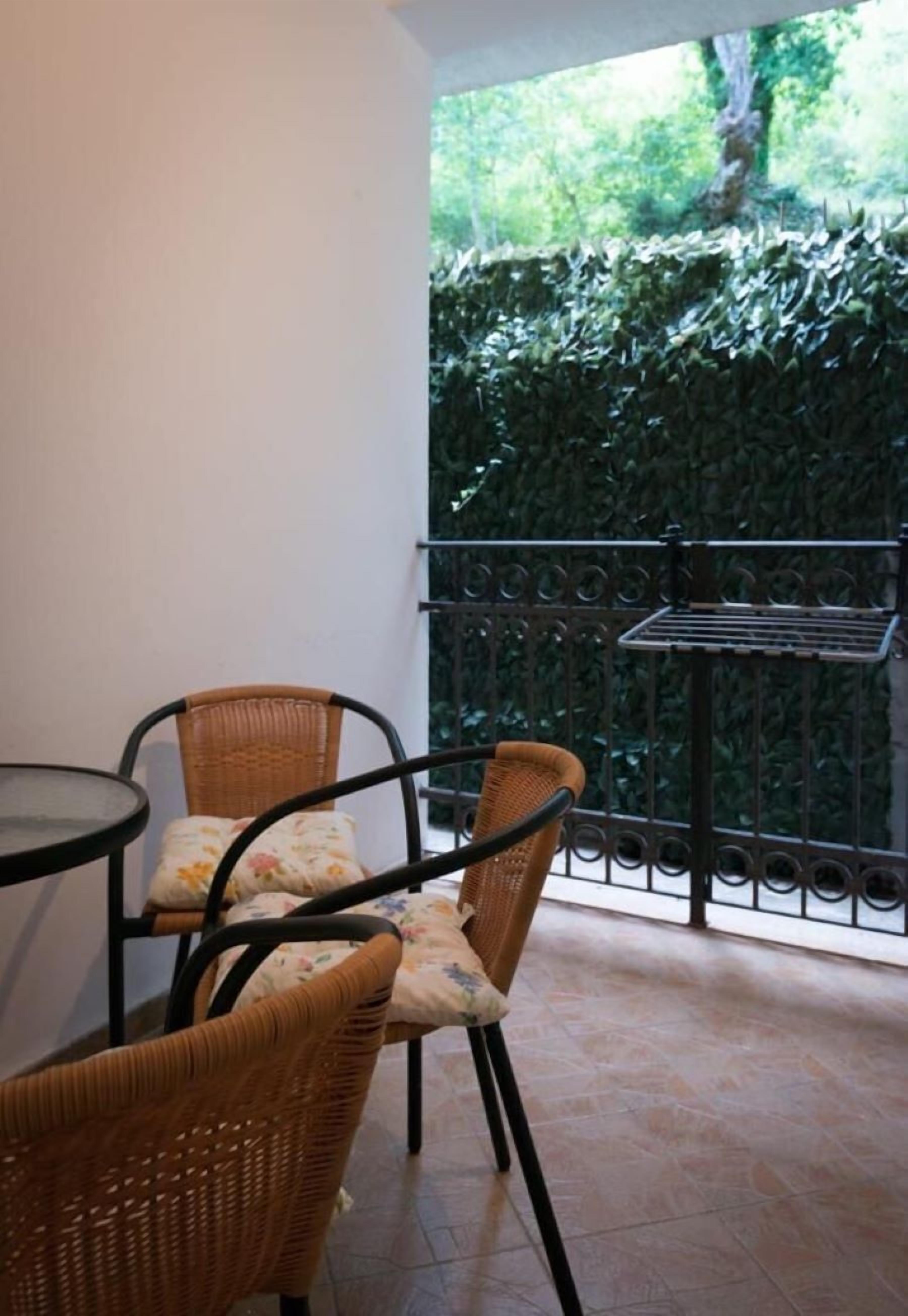 Un balcón con sillones de mimbre, una mesita, barandilla metálica y vista a un seto verde.