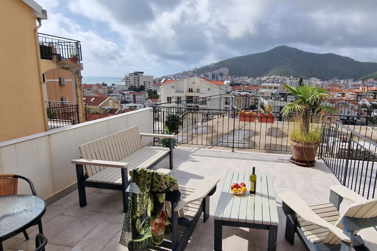 A balcony with outdoor furniture, a table with fruits and a bottle, and a view of a cityscape and mountains.
