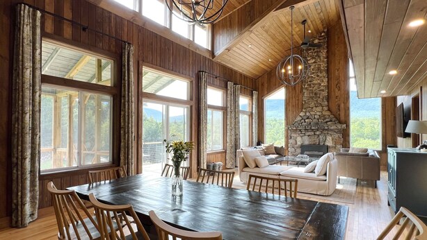 A spacious dining area with a large wooden table and chairs, a stone fireplace, and large windows offering a view of the outdoors. - Spectacular Views, Private River Access Home (Wilmington)