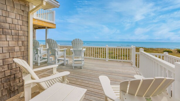 Una terraza de madera con sillas Adirondack blancas y una mesa, con vista a una playa.