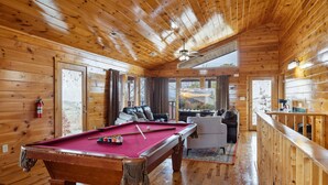 A wooden interior with a pool table, a sofa, and a view through large windows.
