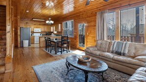 A spacious living area with a leather sofa, a coffee table, and a dining table with chairs. A kitchen with wooden cabinets and a refrigerator is visible in the background.