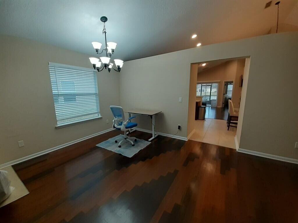 A spacious room with a large window, a desk with a chair, and a chandelier.