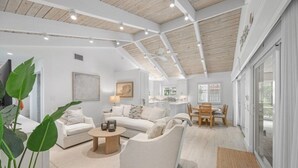 Ein großzügiger Wohnbereich mit hoher Decke, weißen Wänden und Holzboden. Es gibt ein Sofa, einen Couchtisch und einen Esstisch mit Stühlen.