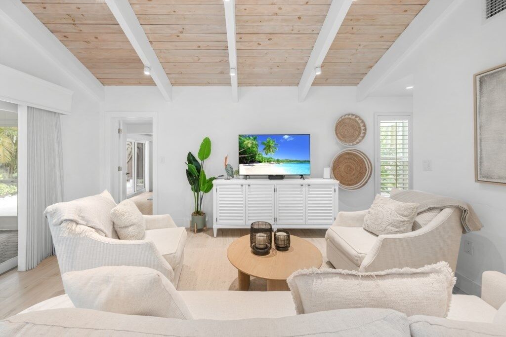 A modern living room with a wooden ceiling, white walls, and a large flat-screen TV on a white cabinet. There are two white sofas and a round coffee table with decorative items.