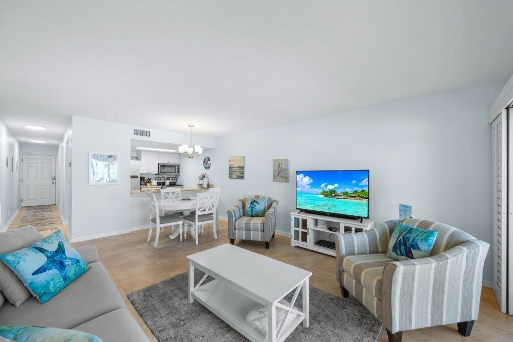 En moderne stue med sofa, lænestol, sofabord og fjernsyn. I baggrunden er der et spiseområde med et bord og stole.