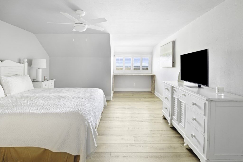 Un dormitorio con cama, mesita de noche, lámpara, cómoda, televisor y ventana con vistas.