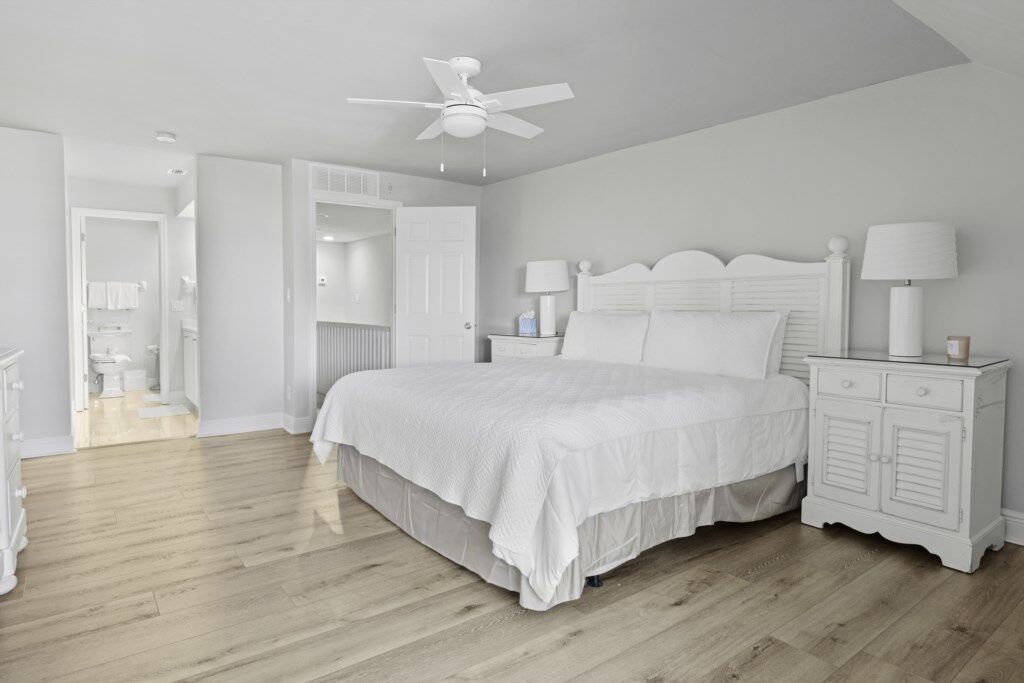 A bedroom with a white bed, a nightstand with a lamp, and a ceiling fan.