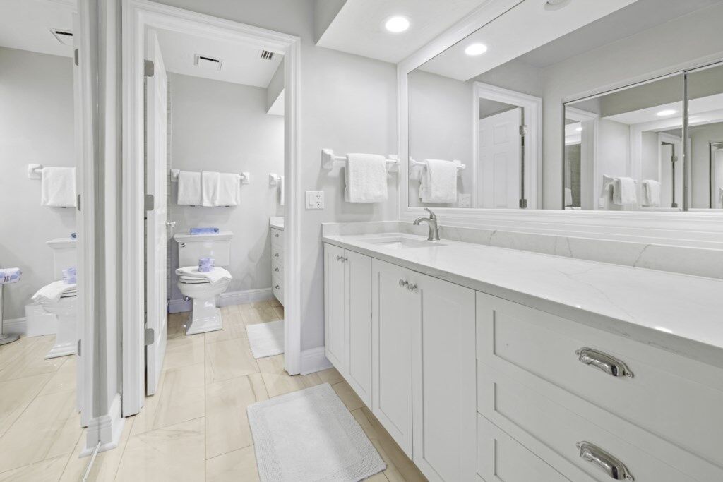 Un baño moderno con un espejo grande, muebles blancos y un inodoro visible al fondo.