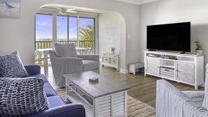 Un salon comprenant un grand téléviseur, un canapé, un fauteuil et une petite table. Il y a également un balcon avec vue sur l’océan.