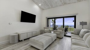 A spacious living room with a large window offering a view of the ocean, a flat-screen TV mounted on the wall, and a seating area with a sofa and ottomans. - South Seas Beach Villa 2535 4 Bedroom Home by RedAwning (Captiva)