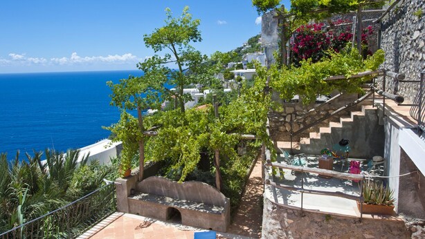 A coastal building with a stone staircase, a small garden with potted plants, and a view of the sea.
