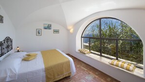 A bedroom with a large arched window, a bed with white linens and a yellow throw, and a brick floor.