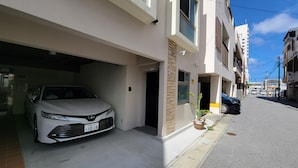 A modern building with a white facade, a car parked in the garage, and a small potted plant on the sidewalk. - Villa Feliz 101 (Naha)