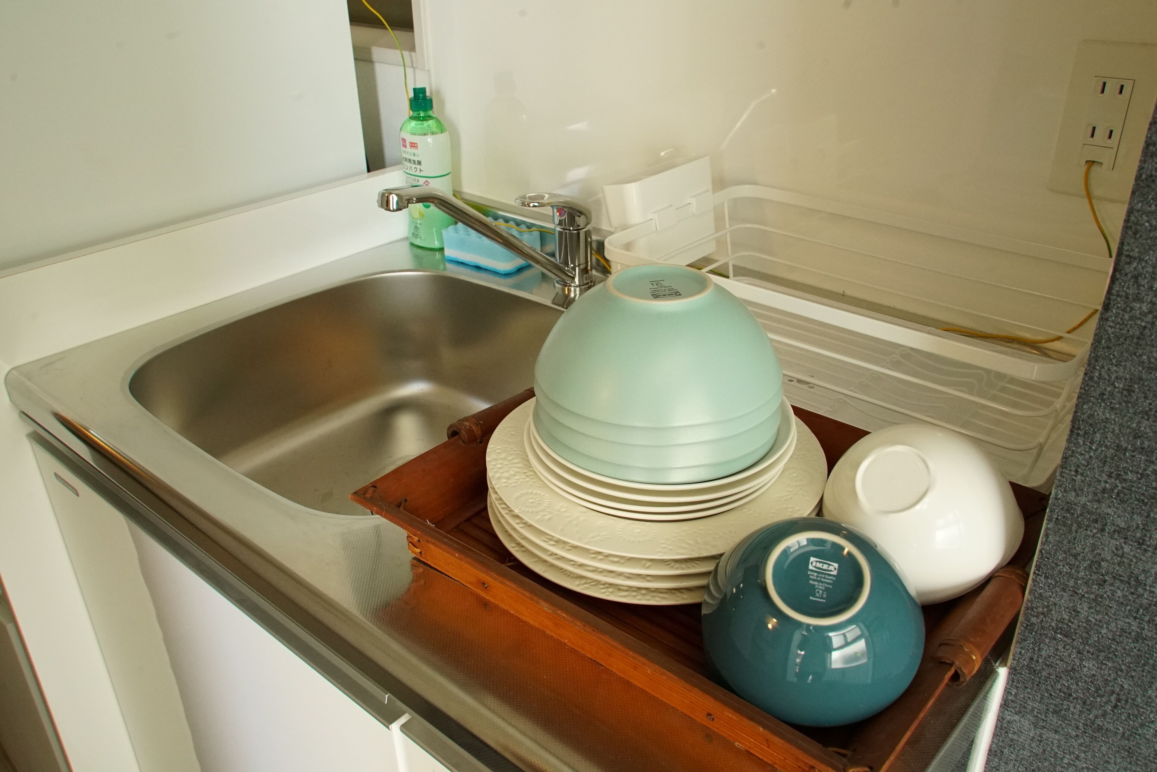 A kitchen sink with a stainless steel basin, a wooden tray holding stacked plates and bowls, and a green bottle of dish soap.
