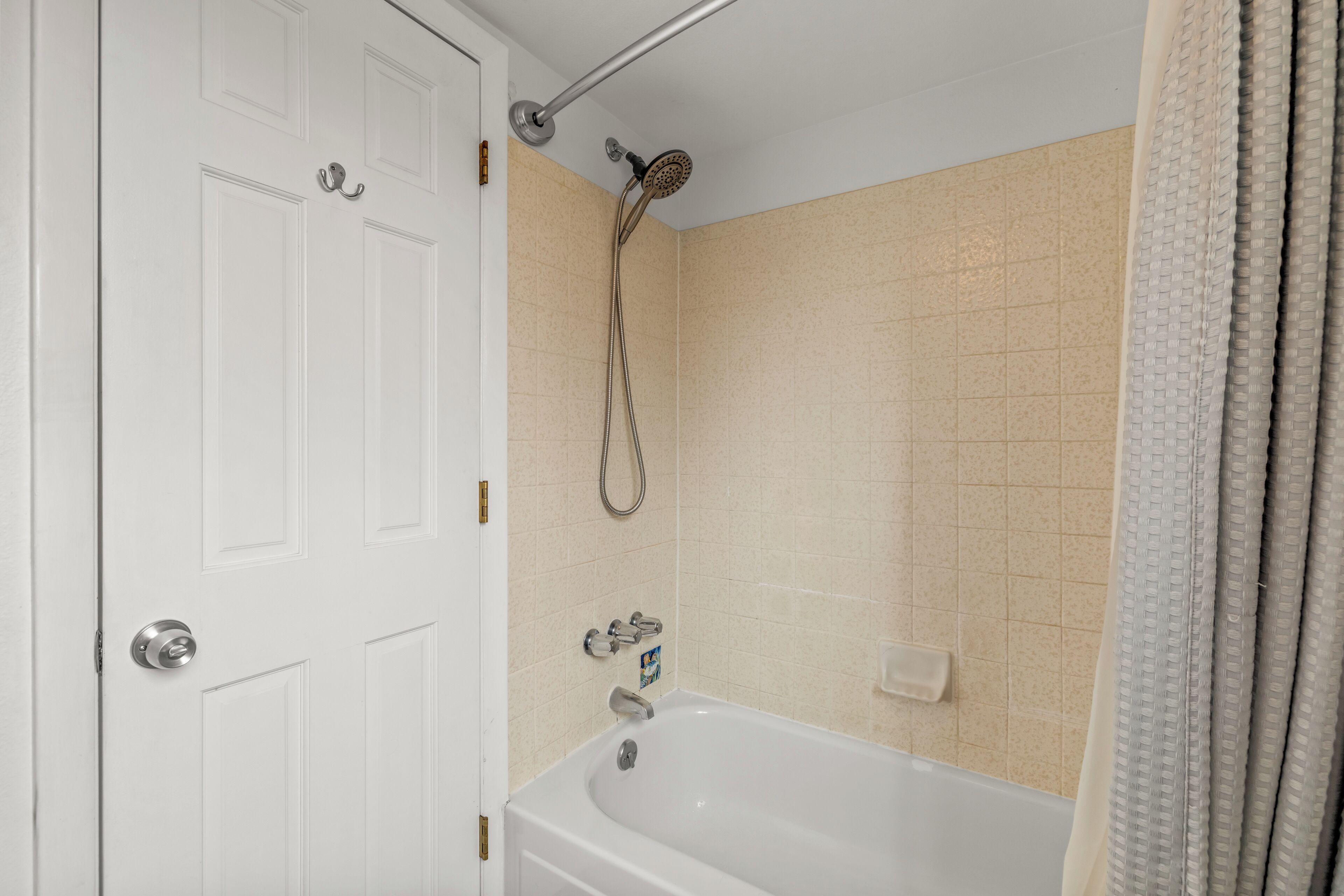 Un bagno con una porta bianca, una vasca, un soffione e pareti rivestite di piastrelle color crema.