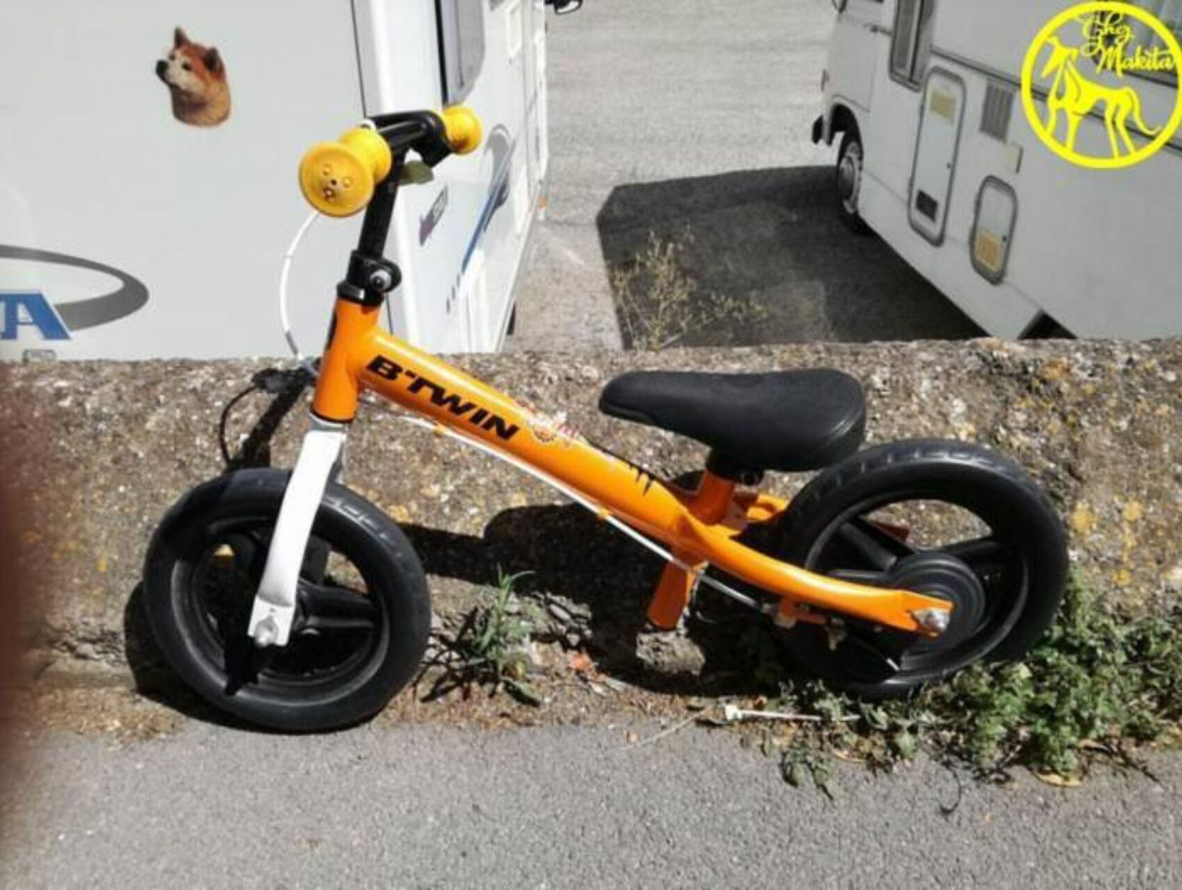 A yellow children's bicycle with the brand name 'B'TWIN' on the frame, parked on a concrete surface next to a white vehicle.