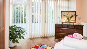 A room with a large window covered by white blinds, a wooden dresser, a framed picture, a vase with a plant, a colorful rug, and a white towel with a pink flower. - Abbey View Farm Cottage (Uttoxeter)