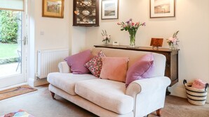 A cozy living room with a beige sofa, a wooden cabinet, and a fireplace. There are framed pictures on the wall and a vase with flowers on the mantel. - Abbey View Farm Cottage (Uttoxeter)