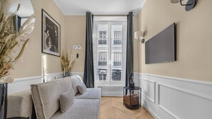 Una sala de estar moderna con un sofá, un televisor de pantalla plana, un jarrón con plantas secas y vistas a un edificio a través de una ventana.