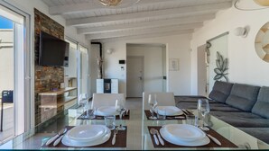 Un espace repas moderne avec une table en verre pour deux personnes, un canapé gris et un téléviseur fixé à un mur en briques.