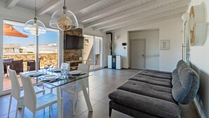 Un salon moderne avec une table en verre, des chaises blanches et un canapé gris. On y trouve également un coin cuisine avec un mur arrière en pierre et une grande fenêtre.