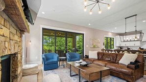 A spacious living room with a fireplace, a brown leather sofa, two blue armchairs, a coffee table, and a large window offering a view of greenery.
