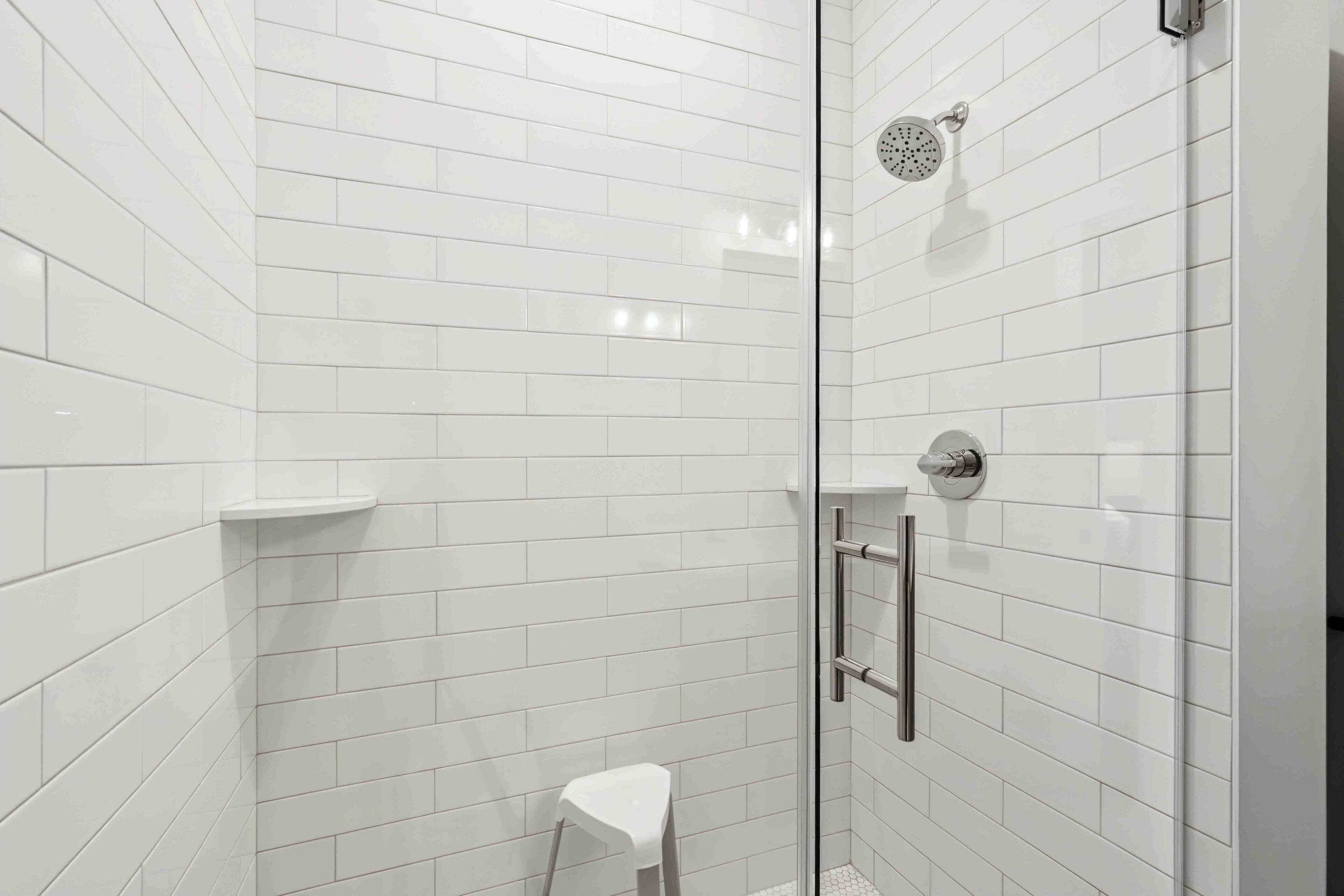 Um chuveiro moderno com porta de vidro, paredes azulejadas em branco e um banquinho branco pequeno.