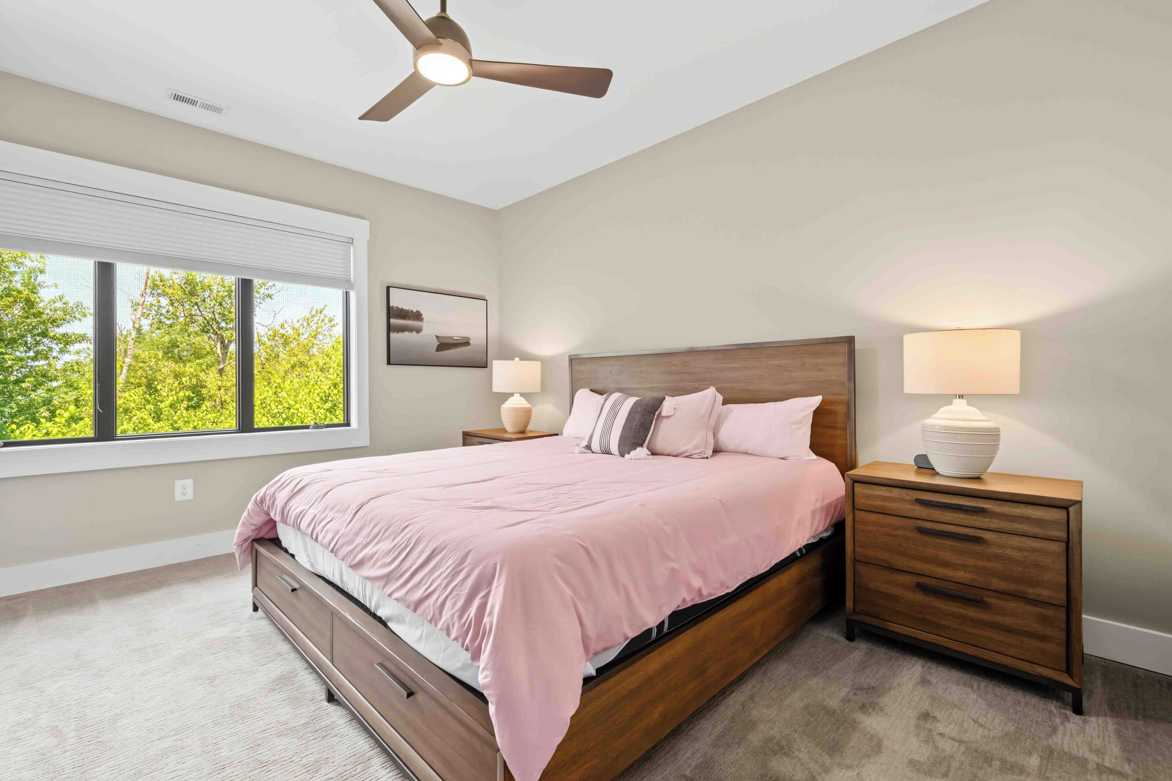 A bedroom with a bed, nightstands, a ceiling fan, and a window with a view of trees.