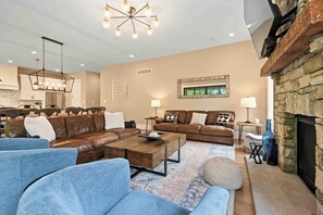 A spacious living room with a large brown leather sofa, a wooden coffee table, and a blue armchair. There is a stone fireplace and a chandelier. - Mountain Mindset by Taylor Made Deep Creek (McHenry)
