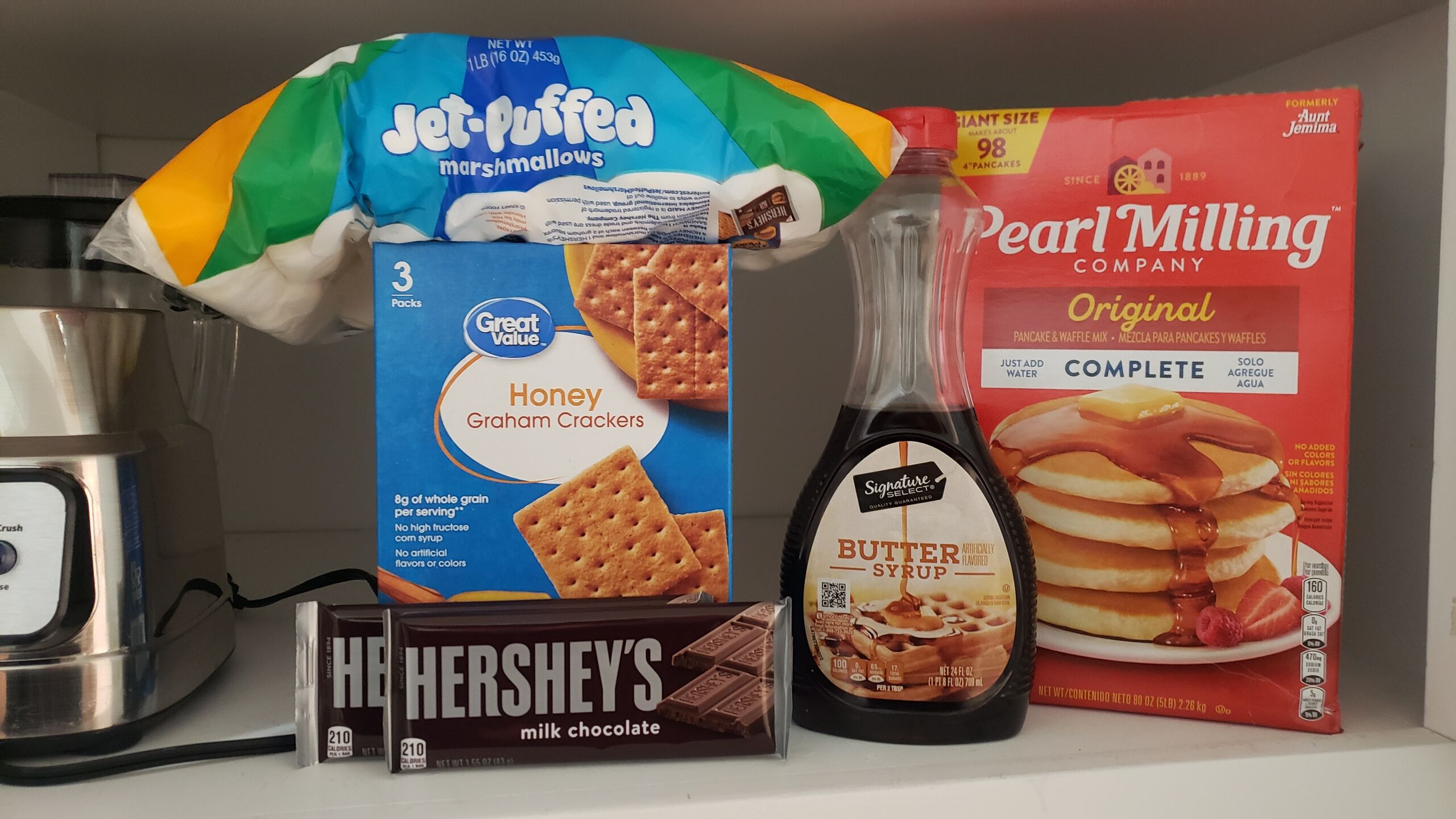 A kitchen shelf with various food items including Jet-Puffed marshmallows, Great Value Honey Graham Crackers, Pearl Milling Original pancake mix, Hershey's milk chocolate, and a bottle of butter syrup.