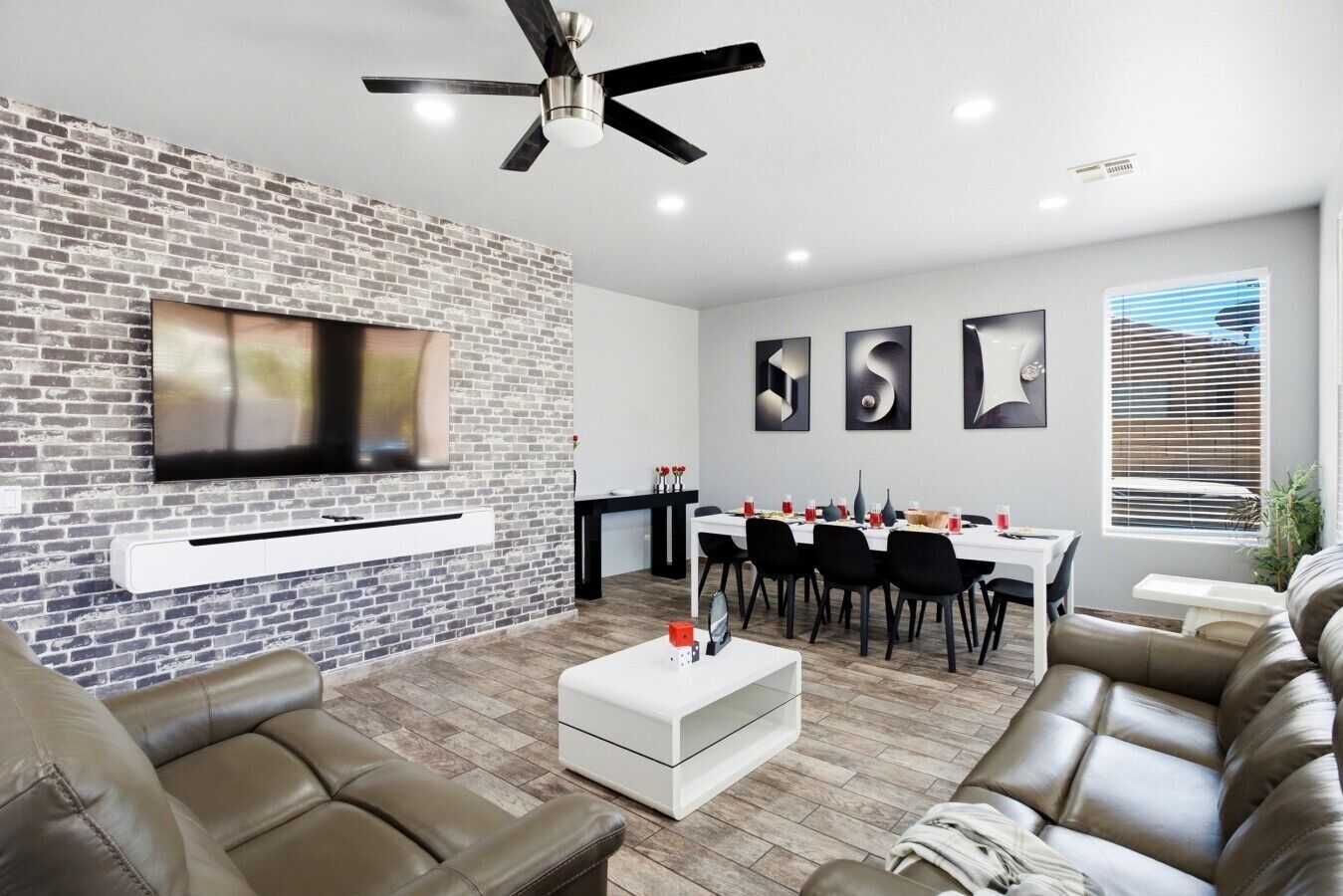 Uma sala de estar moderna com uma parede de destaque em tijolo, uma TV de tela plana, sofás de couro, uma área de jantar com mesa branca e um ventilador de teto.