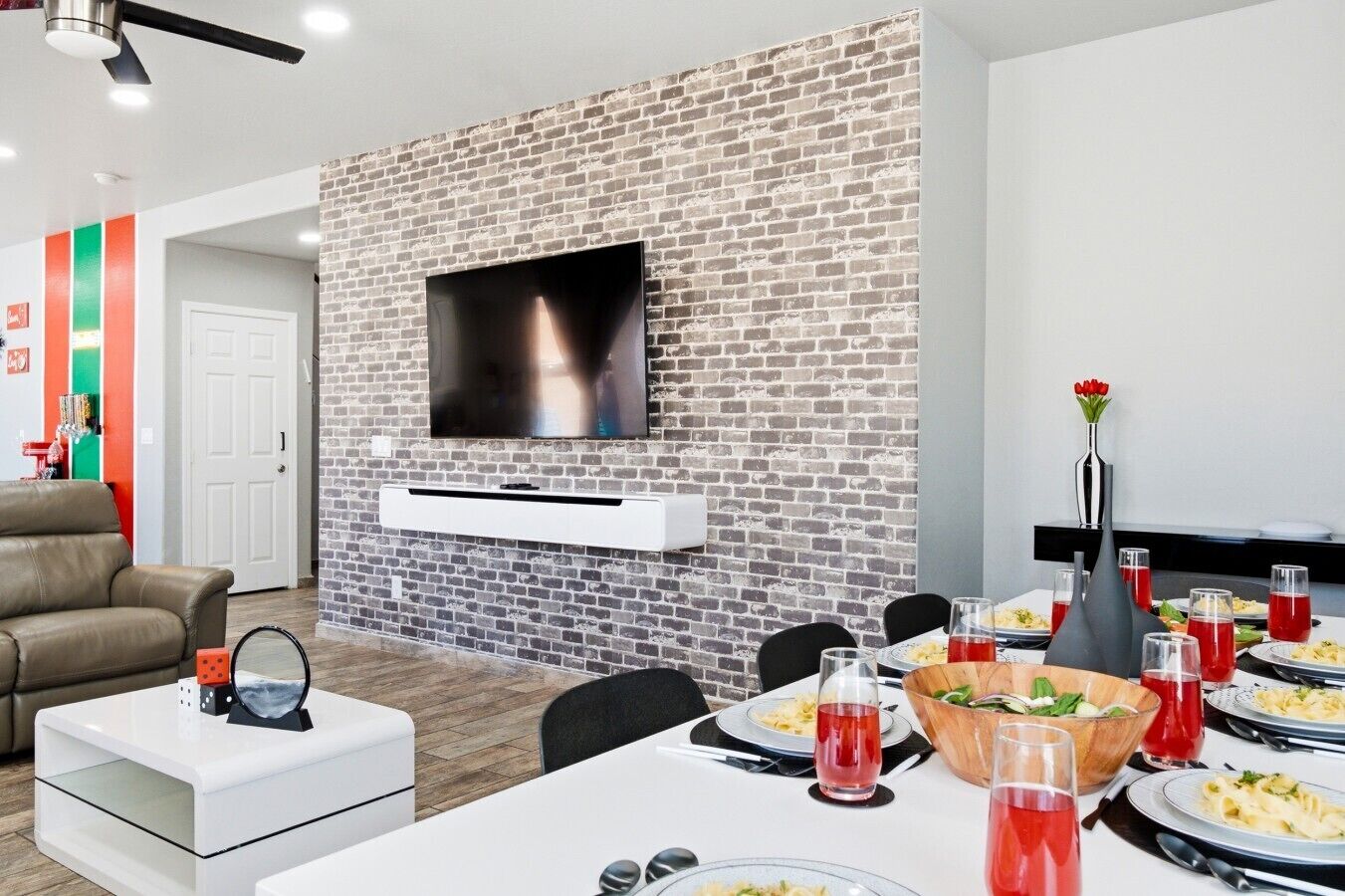 A modern living room with a brick wall, a flat-screen TV, a dining area with a white table set for a meal, and a sofa.